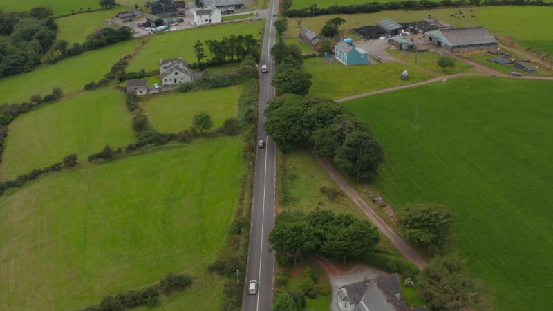 High angle view of cars driving on straight road leading around farms. Tilt up reveal panoramic view of countryside with fields and pastures. Ireland