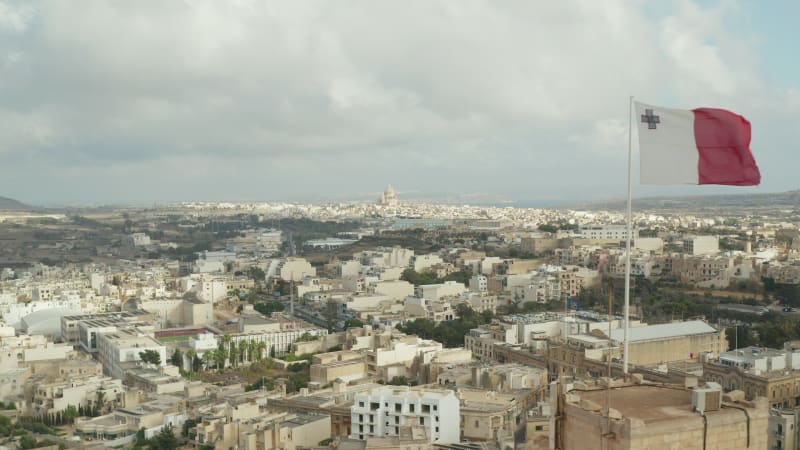 Slow Establishing Shot passing by Flag of Malta waving in revealing City on Gozo Island with cloudy Blue Sky , Aerial forward