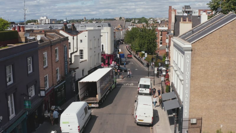 Fly over streets in urban neighbourhood. Pedestrians walking along parked vans and cars. Truck unloading. London, UK