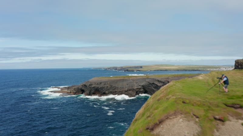 Photographer standing on cliff edge and taking pictures. Revealing waves crashing on rocky coast. Panoramic view of shore. Kilkee Cliff Walk, Ireland