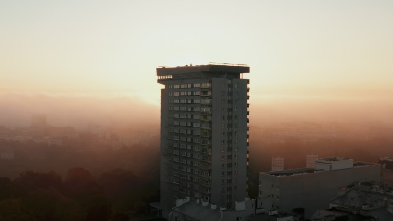 Fly over standalone tall apartment house in town. Revealing rising sun in mist. Morning shot through sparse fog or low clouds. Warsaw, Poland