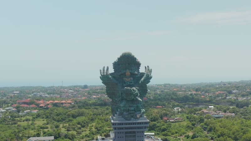 Giant copper statue of Garuda Wisnu Kencana in cultural park in Bali, Indonesia. Aerial dolly retreating view of large religious sculpture rising above the city