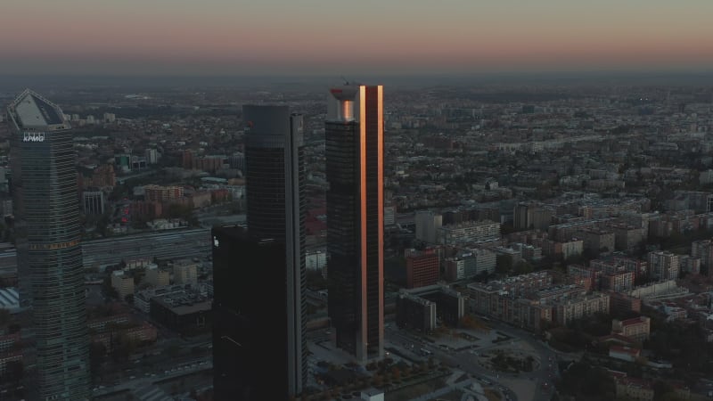 Descending aerial footage of futuristic tall skyscrapers in Cuatro Torres Business area at dusk. Glossy facade reflecting colourful twilight sky.