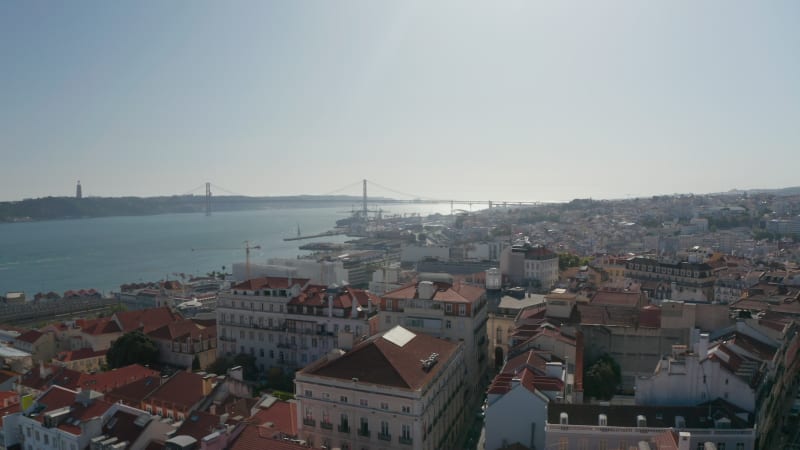 Panoramic aerial view of town with long cable-stayed bridge spanning Tagus river. Drone camera descending between buildings and gradually hiding view. Lisbon, capital of Portugal.