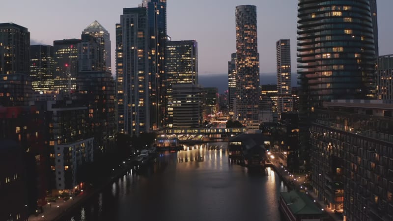 Forwards fly above water surface in modern business district in evening. Lights reflecting on water. London, UK