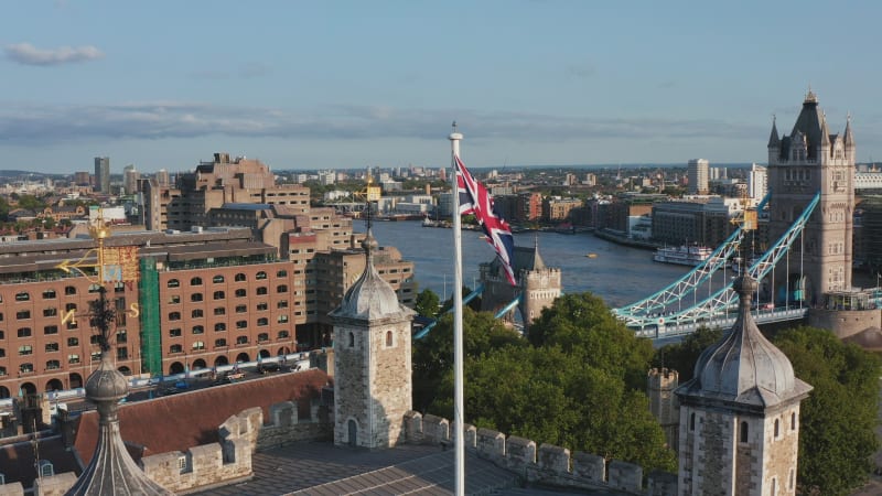 Union Jack slightly waving in breeze. Backwards reveal of white Tower and rest of Tower of London castle complex. Tower Bridge across River Thames in background. London, UK