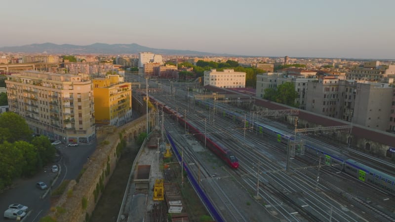 Passenger trains driving on multitrack railway lines in city. Multistorey residential buildings lit by evening sun. Rome, Italy