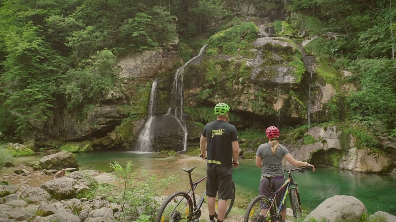 Aerial view of cyclists standing with their bicycles in front of a waterfall.