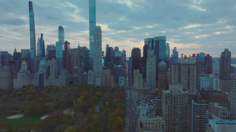 Forwards fly along autumn Central park at dusk. Aerial footage of high rise buildings in downtown surrounding park. Manhattan, New York City, USA