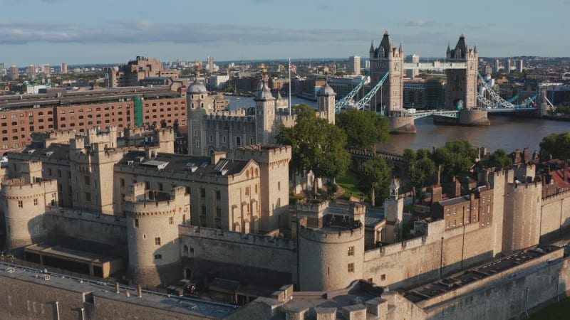 Forwards fly above Tower of London in golden hour. Impressive stone fortification of medieval castle. Old White Tower with four corner towers and Union Jack on pole. London, UK