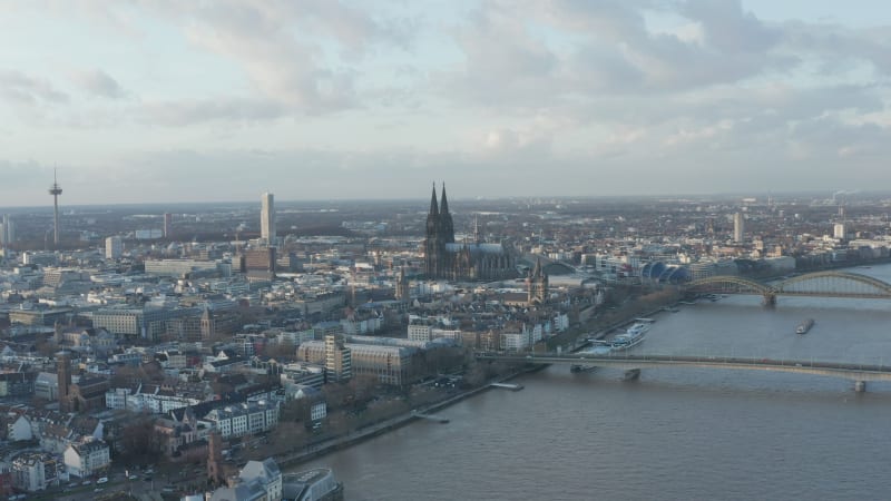 Backwards fly above Rhine river in city at dusk. Busy bridges over rippled water surface. Gothic Kolner Dom towering above surrounding development. Cologne, Germany