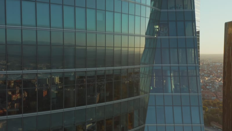 Ascending footage along windows and glass wall of office skyscraper reflecting buildings in town and setting sun. Revealing Spanish flag on rooftop.