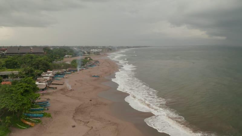 People relaxing on a tropical sand beach before the storm in Canggu, Bali. Aerial dolly view of colorful boats  and tourists on famous tourist beach in Indonesia