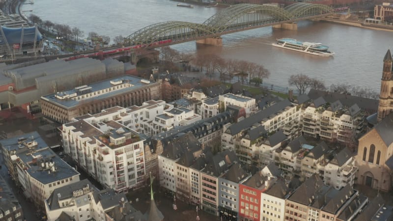 Fly above urban borough. Tilt up reveal commuter train passing over wide Rhine river on bridge. Cologne, Germany