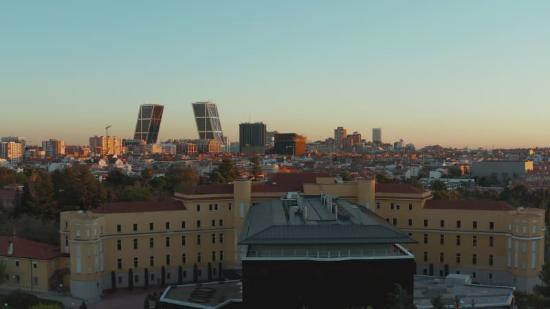 Fly over historic building with modern extension. Revealing cityscape with business skyscrapers lit by setting sun.