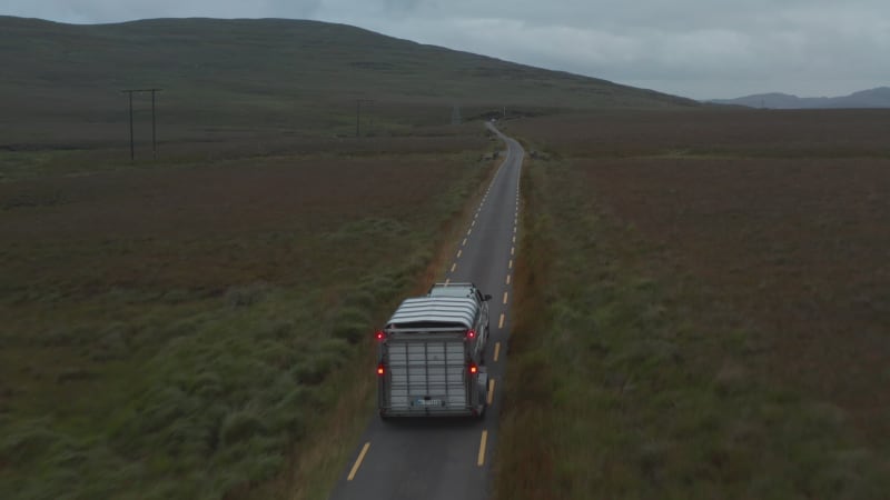 Forwards tracking of offroad car with horse trailer driving on narrow road in countryside. Pastures and grasslands along road. Ireland