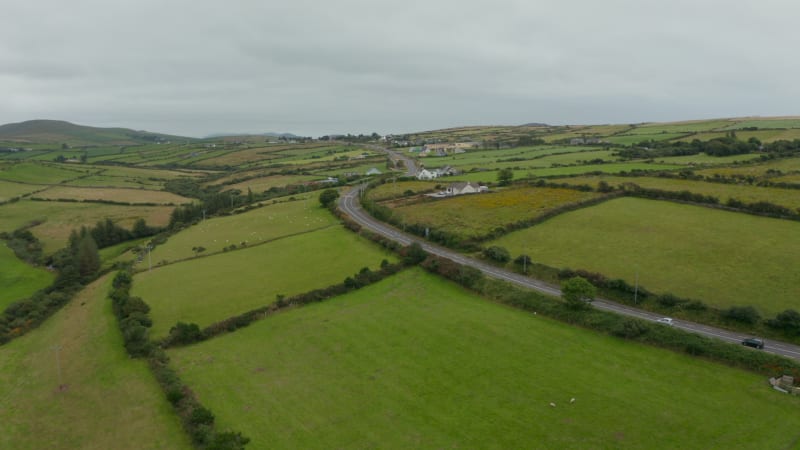 Forwards fly above cars driving on road between green meadows and pastures. Cloudy day in countryside. Ireland