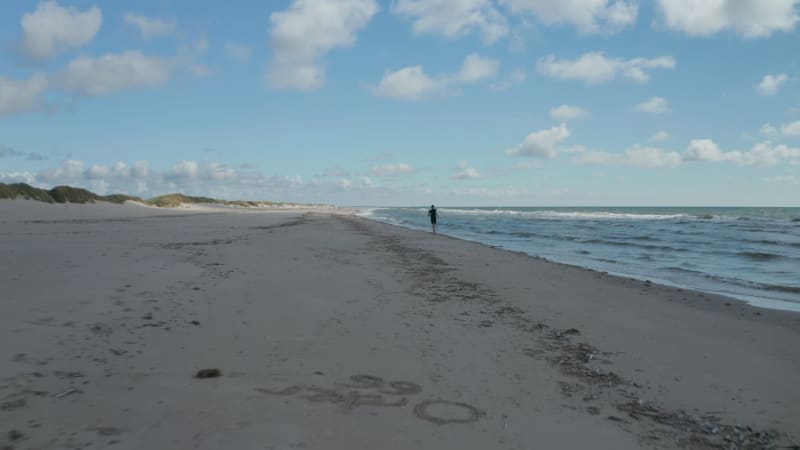 Forwards tracking of person running on beach. Sands in cloud shadow. Waves washing sea coast. Denmark