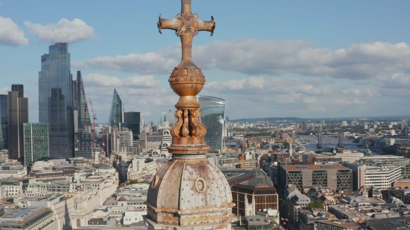 Panorama curve shot footage of decorative cross on top of church tower with cityscape in background. Group of tall modern skyscrapers in City business district. London, UK