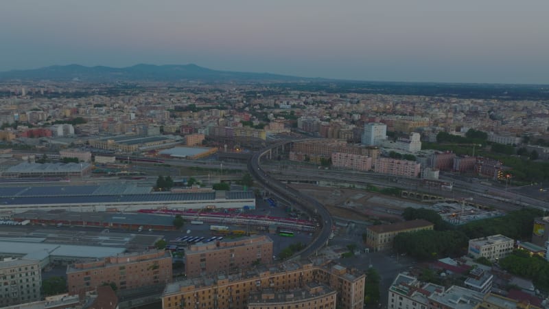 Forwards fly above large city after sunset. Group of modern train units parking in depot. Elevated road bridge over railway tracks. Rome, Italy