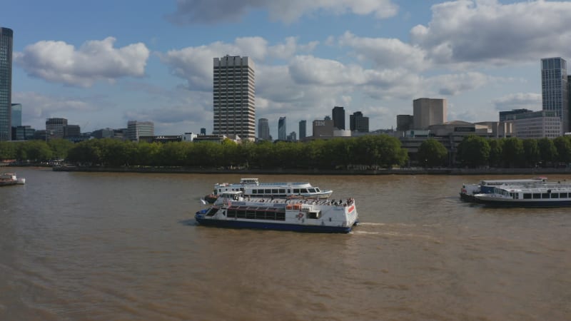 Tracking shot of cruise ship floating on wide water surface of River Thames. One Blackfriars and skyscrapers in City financial hub in background. London, UK