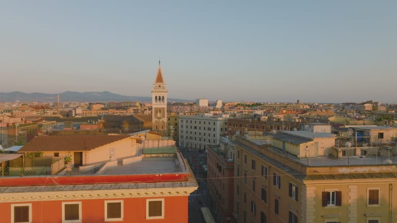 Aerial footage of old apartment buildings in urban borough at golden hour. Tower with decorations protruding above town development. Rome, Italy