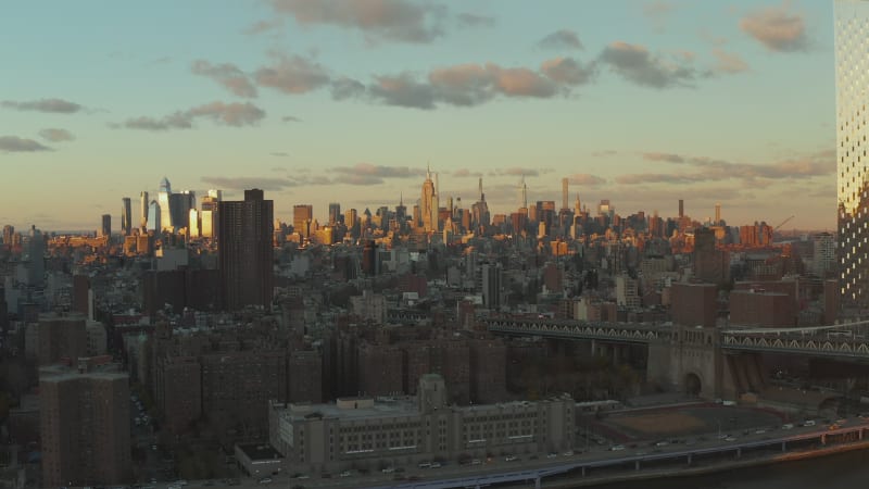 Sliding reveal of waterfront and bridges over river. Cityscape with tall skyscrapers lit by evening sun. Manhattan, New York City, USA