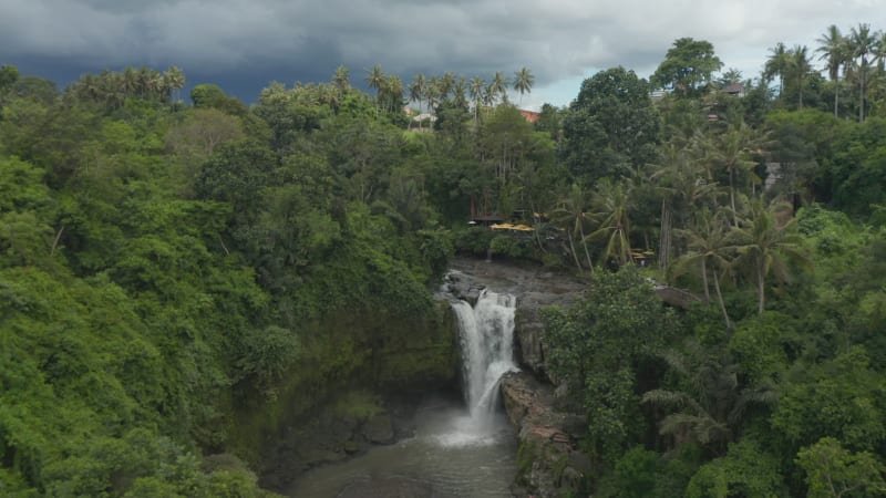 Tilting aerial view of a stunning waterfall in the jungle with beautiful outdoor restaurant at the top of the cliff in Bali rainforest