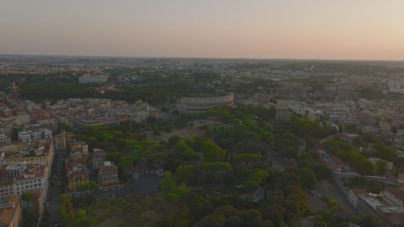 Forwards fly above Oppian Hill park with historic landmarks. Heading towards ancient Colosseum amphitheatre. Panoramic view of city at dusk. Rome, Italy