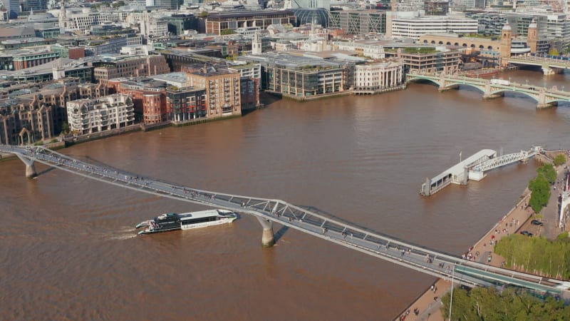 Fly above Thames River at Millennium footbridge. Tilt up reveal of modern tall office buildings in City  business hub. London, UK