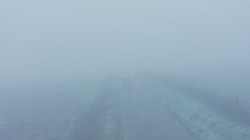 Aerial drone video of car driving in misty thick foggy, bad and dangerous driving conditions on a rural countryside road, 4 wheel drive with trailer driving past fields in England in mist and fog