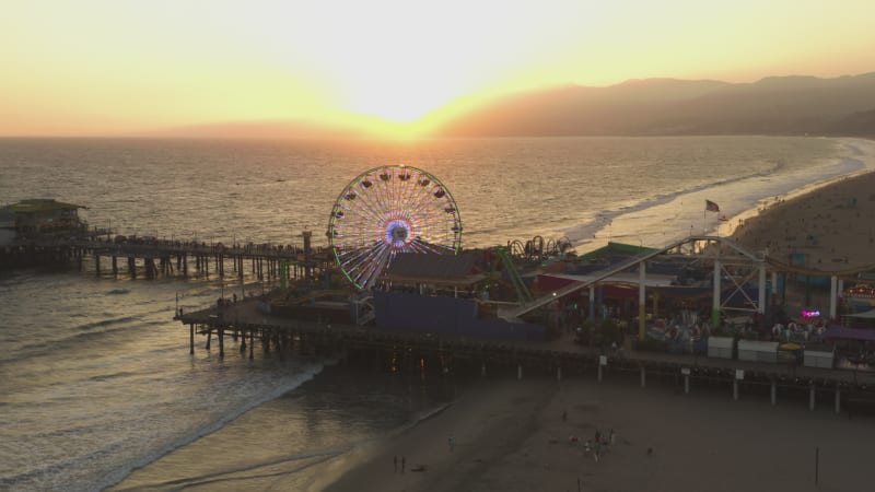 Coming down on Santa Monica Pier Ferris Wheel, Los Angeles at beautiful Sunset with Tourists, Pedestrians walking having fun at theme park rollercoaster with ocean view waves crashing