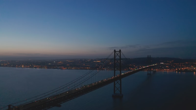 Night aerial view of 25th of April Bridge connecting Lisbon and Almada. Car headlights on cable-stayed highway bridge over Tegus river. Drone flying forwards. Lisbon, capital of Portugal.