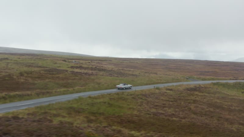 Tracking of single vintage convertible car with textile roof driving on road in countryside on cloudy day. Ireland