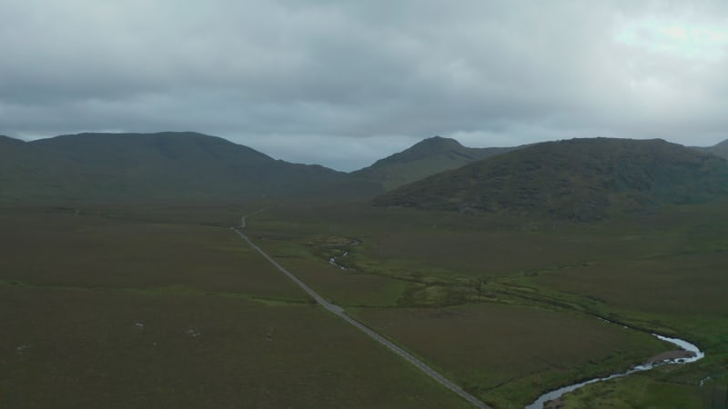 Aerial panoramic footage of landscape with mountains in background. Stream winding along straight road. Ireland