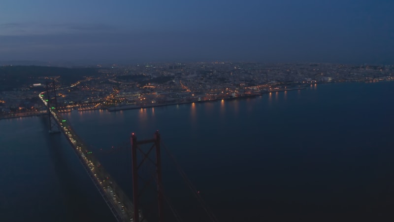 Night panoramic aerial view of town on rivershore. Light of cars driving on bridge and city street light in downtown. Footage from flying drone. Lisbon, capital of Portugal.