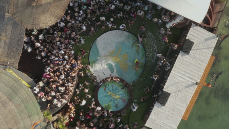 People dancing and partying at tropical Beach club Festival Party by the ocean in sunny weather with Palm trees and Pools, Aerial Birds Eye View Top Shot from above