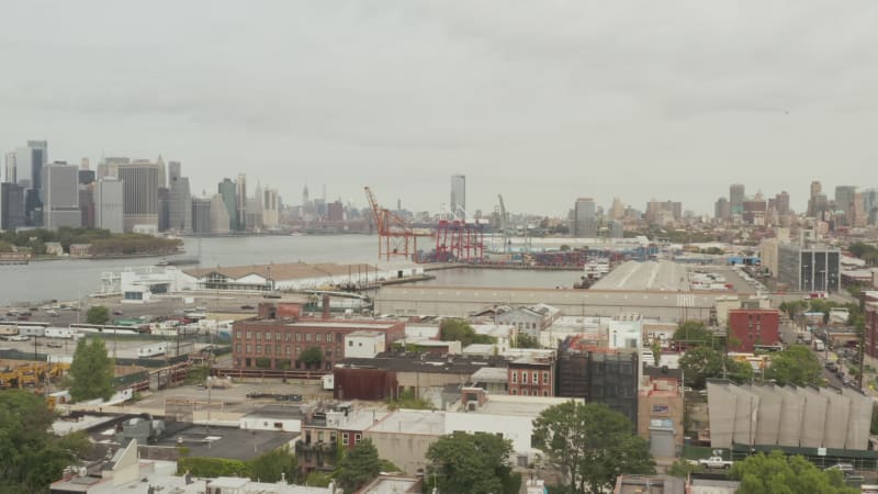 Over NYC Docks Warehouses towards industrial cranes on cloudy day with view of Manhattan Skyline in