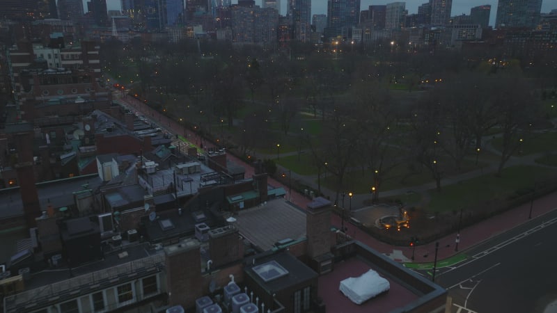 High angle view of cars driving on road along park. Tilt up reveal cityscape with modern high rise buildings at dusk. Boston, USA