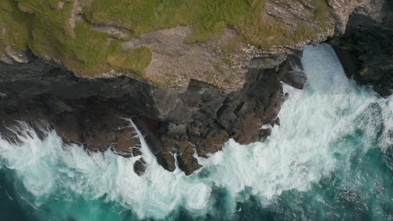 Aerial birds eye overhead top down ascending view of rough waves crashing on rocky coast. Green grass on top of cliffs high above clear water. Kilkee Cliff Walk, Ireland