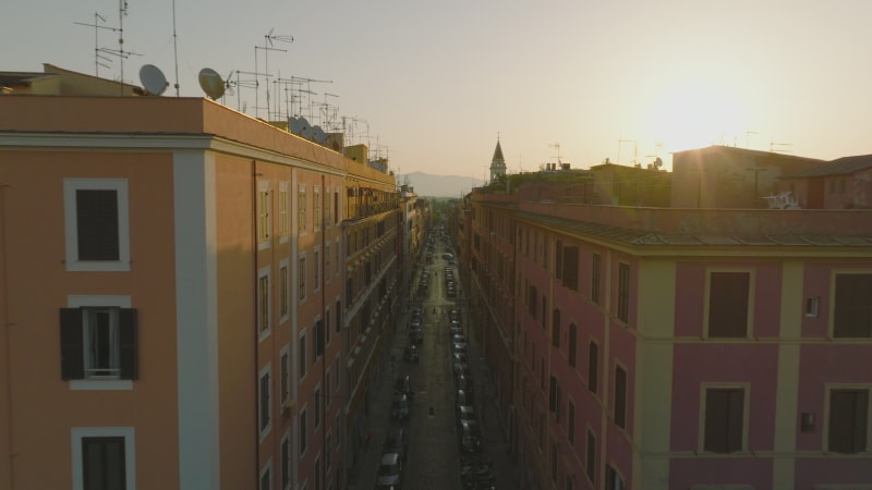 Forwards fly along top of apartment houses in street of urban borough at sunrise. Revealing silhouette of tower against bright sky. Rome, Italy