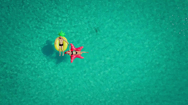 Aerial view of man and woman floating on inflatable mattresses holding hands.