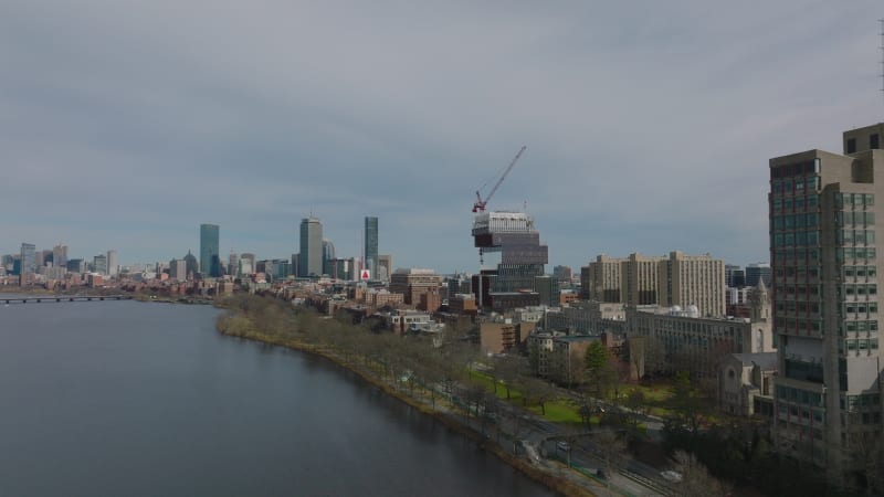 Forwards fly along waterfront. Construction of modern university building. Downtown skyscrapers in background. Boston, USA