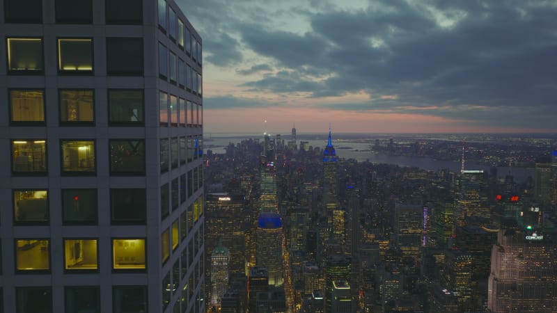 Tight fly above top storeys of modern skyscraper towering above city. Reveal panoramic shot of downtown at dusk. Manhattan, New York City, USA