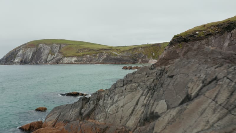 Fly over coastal rocks. Waves washing jagged sea coast. Revealing people on small sand beach under cliff. Amazing nature scenery. Ireland