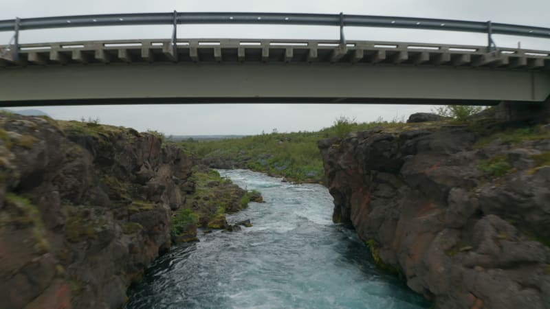 Aerial view drone flying under bridge over flowing river in rocky countryside in Iceland. Birds eye spectacular icelandic highlands with river powerfully flowing