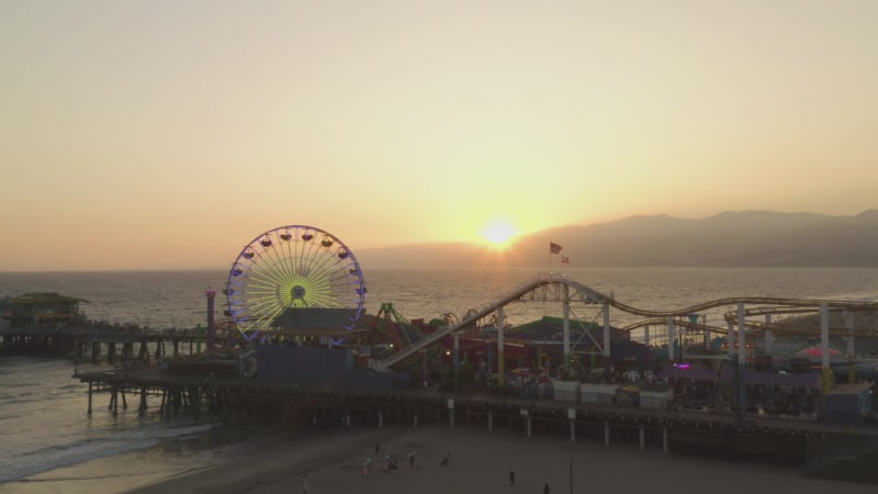 Flying towards Santa Monica Pier, Los Angeles at beautiful Sunset with Tourists, Pedestrians having fun at Ferris Wheel with ocean view waves crashing, Drone Perspective