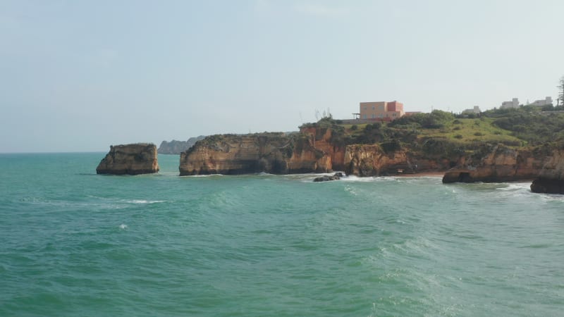 Aerial drone view of waves crashing against colorful Lagos cliff coastline, drone flying foward above rocky coast reveal amazing sea landscape, day