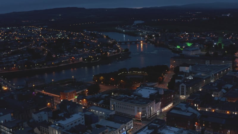 Fly above city in evening. Aerial view of river flowing through night town with street lights. Limerick, Ireland