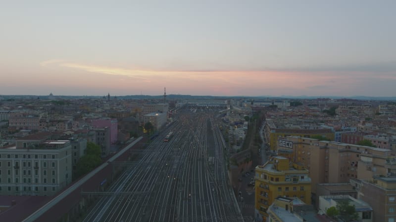 Aerial slide and pan footage of wide railway body with multiple tracks leading through city. Roma Termini train station at dusk. Rome, Italy